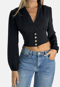 Zwarte wikkelblouse met lange pofmouwen, V-halsontwerp en vijf houten knopen aan de voorkant, gecombineerd met lichtblauwe denim jeans.