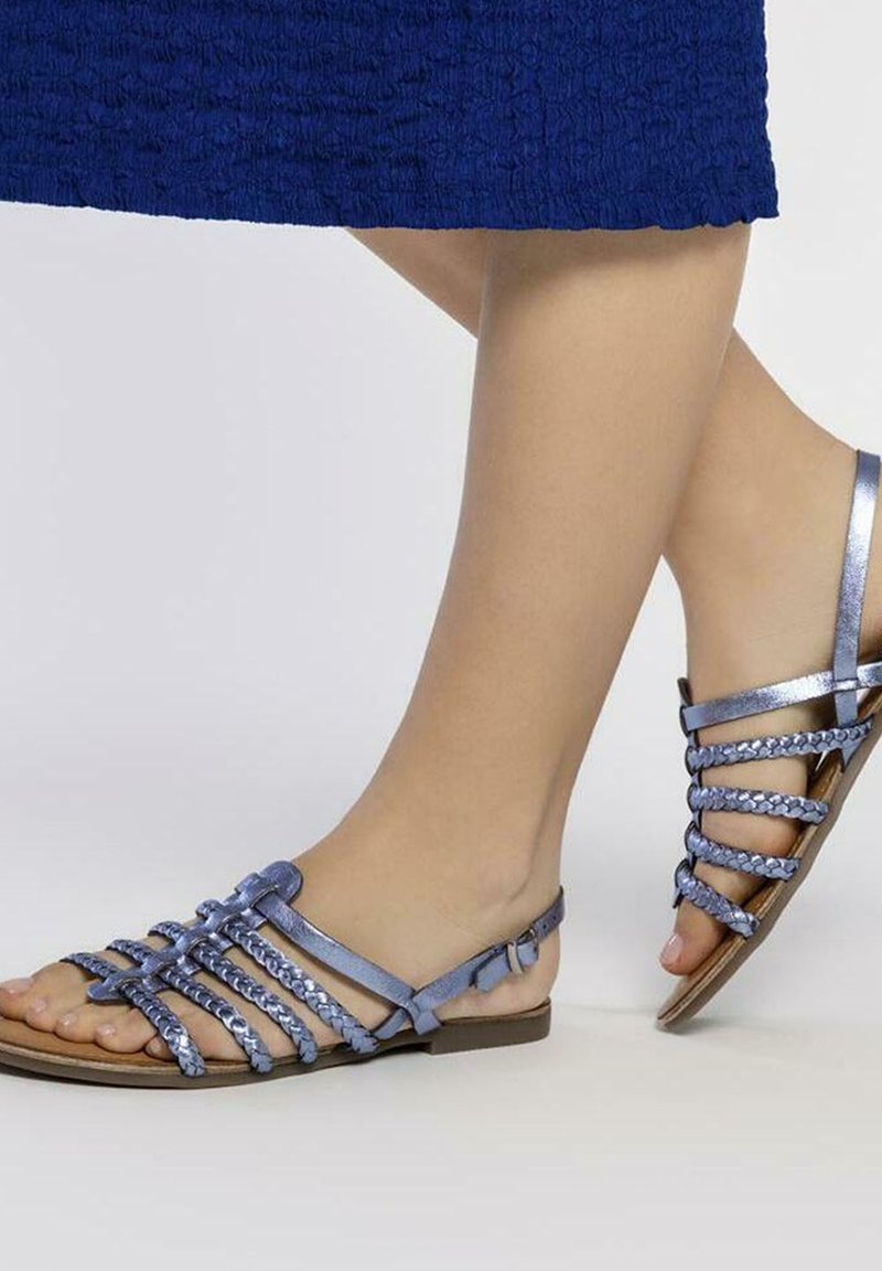 Sandalias de tiras en material metálico azul claro, con detalles trenzados, diseño de punta abierta, suela plana y correa en el tobillo ajustable.