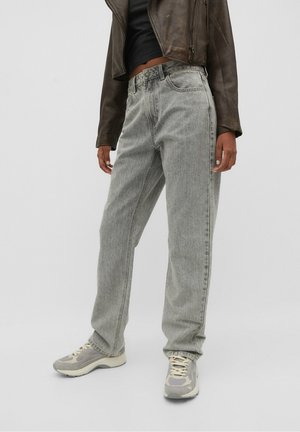 Person wearing light gray denim jeans, beige and gray sneakers, black cropped top, and dark brown leather jacket against a plain background.