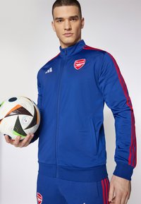 Blaue Sportjacke mit roten Streifen, versehen mit einem Arsenal-Logo und Reißverschluss vorne. Das Modell hält einen gemusterten Fußball.