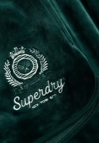 Tissu vert foncé avec un logo brodé blanc comprenant une couronne, une couronne de laurier et le texte "Superdry New York City".
