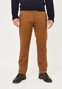 Braune Baumwollhosen mit geradem Bein, Fronttaschen und kombiniert mit schwarzen Lederschuhen. Die Textur ist glatt und leicht matt.