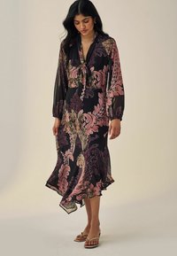 Rochie neagră cu flori, cu mâneci lungi, din material translucid, detaliu cu cravată la gât și o fustă floștită cu un hem hi-low. Are modele cu frunze roz și verzi.