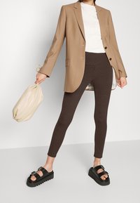 Leggings marrón con estampado, blazer ligero beige, camiseta blanca, bolsa beige y sandalias negras gruesas con hebillas.