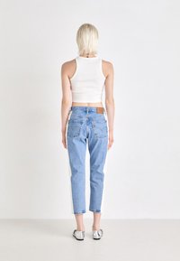 Biely krátky top bez rukávov s modrými džínami, ktoré majú biele bočné panely. Modelka stojí s rukami po bokoch, na nohách má strieborné loafers a krátke vlasy.
