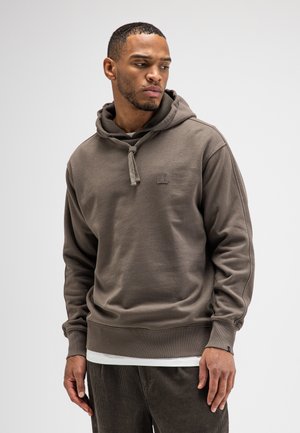 Mann trägt braunen Hoodie mit Kordeln und braune Hose, schaut nach links vor einfachem hellem Hintergrund.