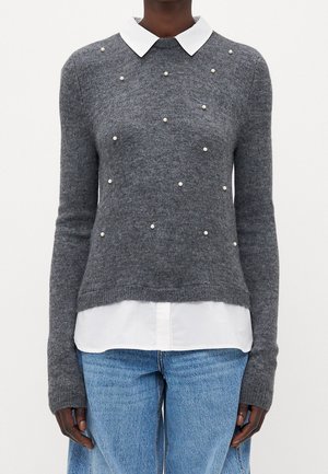 Person iført en grå sweater pyntet med spredte små perler, hvid skjorte med krave under og lyseblå jeans.