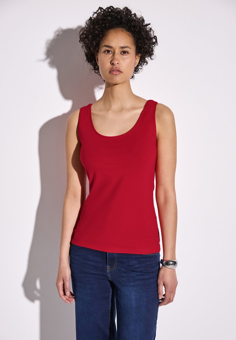 Top rosso in tessuto liscio, con scollatura a barca e design senza maniche. Abbinato a jeans blu scuro.