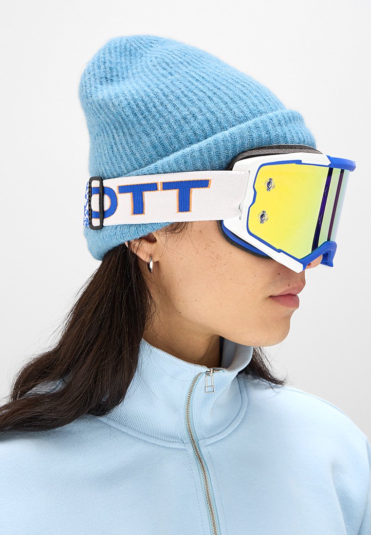 Gafas de esquí con lente reflectante amarilla y acentos azules, combinadas con un gorro de punto azul claro y una chaqueta con cremallera.