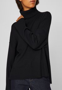 Μαύρο πουλόβερ με ψηλό λαιμό, άνετη εφαρμογή, ριγέ υφή και ελαφρώς χαμηλωμένους ώμους, διαθέτει πλαϊνές σχισμές και άνισο τελείωμα.