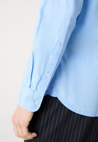 Camicia azzurro chiaro a maniche lunghe con polsino a bottoni, realizzata in un tessuto strutturato, taglio sartoriale e dettaglio con spacco laterale. Abbinata a pantaloni a righe.