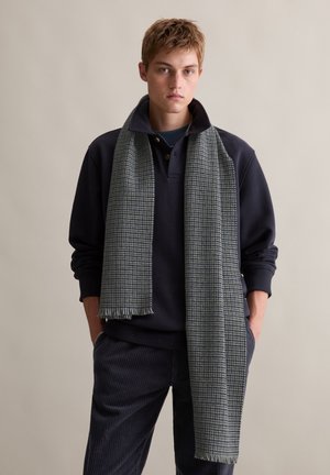 Green and gray checkered scarf draped over a navy pullover, made of soft fabric with fringed edges, styled with corduroy pants.