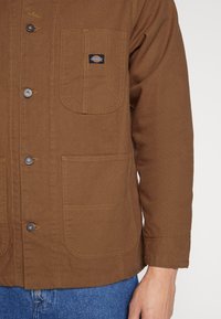 Brown jacket made of durable fabric, featuring a button front, two large pockets, contrasting stitching, and a logo patch on the chest pocket.