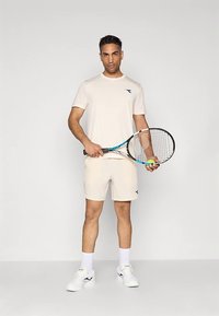 Beige athletic t-shirt and shorts, with a blue logo. The model holds a tennis racket and ball, wearing white sneakers and socks.
