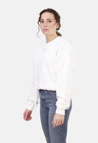 Felpa bianca corta realizzata in misto cotone, con scollo rotondo, maniche lunghe e cordino sull'orlo. Abbinata a jeans blu.