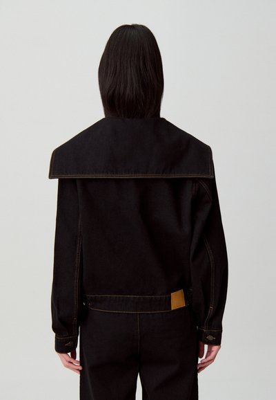 Persona con capelli scuri e lisci che indossa una giacca nera con colletto ampio e pantaloni neri abbinati, vista da dietro su uno sfondo semplice.