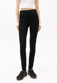 Άτομο που φοράει μαύρο στενό τζιν ψηλόμεσο και μαύρα αθλητικά παπούτσια με λευκές και κόκκινες λεπτομέρειες, στέκεται μπροστά σε λευκό φόντο.