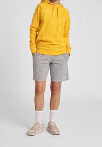 Sudadera con capucha amarilla, pantalones cortos grises y zapatillas beige con detalles blancos. Presenta un logo en la sudadera y bolsillos en los pantalones cortos.