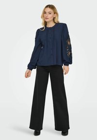 Camicia blu navy con bottoni, maniche lunghe a sbuffo e dettagli in pizzo, abbinata a pantaloni neri a gamba larga e scarpe nere. Tessuto dalla consistenza liscia.