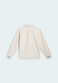 Camisa de manga larga color beige claro con cuello abotonado, detalle de pliegue en la espalda y botones negros en los puños. Tejido suave y texturizado.