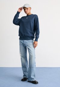 Sweatshirt azul marinho com o logótipo branco da Nike, emparelhado com jeans desgastados de azul claro e sapatos pretos, coberto com um boné cinza claro.