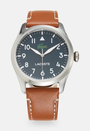 Lacoste laikrodis su tamsiai pilku ciferblatu, dideliais baltais skaičiais ir žaliu krokodilu logotipu. Savybė – nerūdijančio plieno dėžutė ir ruda odinė apyrankė.