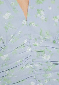 Ljust blå klänning med blomm önster i vitt och grönt. Har en gathered textur och en v-ringad halsringning. Mjukt, lättviktigt tyg.