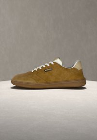 Baskets Versace en daim marron avec lacets blancs, accent beige au talon, et semelle marron ornée d'un motif en relief, présentées sur une surface neutre.