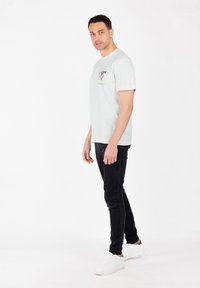 Hombre con cabello corto y oscuro, vistiendo una camiseta gráfica blanca, jeans negros ajustados y zapatillas blancas, de pie contra un fondo blanco.
