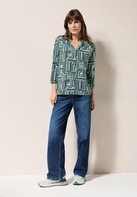 Langarmhemd mit Knopfverschluss in Mintgrün und schwarzem geometrischem Muster. Kombiniert mit lockeren blauen Jeans und hellen Sneakers, vor einem neutralen Hintergrund stehend.