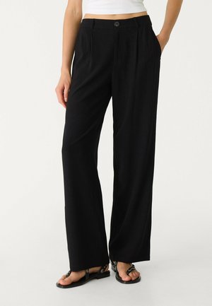 Vrouw draagt zwarte wijde broek met hoge taille en zijzakken, gecombineerd met zwarte platte sandalen en een wit cropped topje.