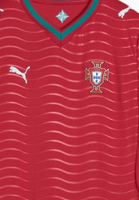 Κόκκινη αθλητική φανέλα με V-λαιμόκοψη, διακοσμημένη με υφή κυματιστού μοτίβου, τυρκουάζ περιμετρικό γιακά, λογότυπο Puma και το εθνικό έμβλημα της Πορτογαλίας.