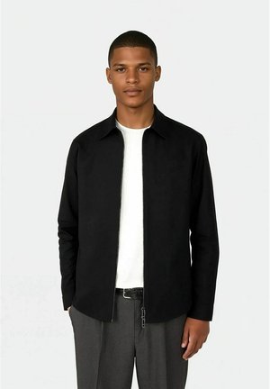 Ung mann iført svart jakke med glidelås over en hvit skjorte og grå bukser, stående foran en ensfarget lys bakgrunn.