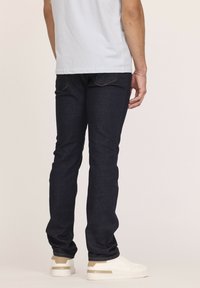 Jean en denim bleu foncé avec une coupe slim, présentant des poches arrière et une texture lisse. Associés à des baskets blanches pour un look décontracté.