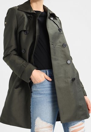 Vrouw die een donkergroene dubbelgeknoopte trenchcoat draagt over een zwart shirt en gescheurde blauwe spijkerbroek, één hand in de zak.
