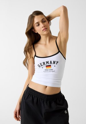 Jeune femme ajustant ses cheveux, portant un débardeur blanc avec le texte "Germany" et le drapeau, ainsi qu'un pantalon noir à taille élastique sur un fond uni.