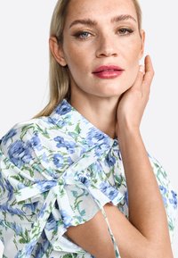 Short-sleeved blouse with blue and green floral pattern on white fabric; features a pointed collar and tied accent at the sleeves.