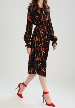 Kvinde gående i en sort midi-kjole med røde og orange blomsterprint, iført røde højhælede ankelremssko.