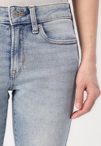Jeans de mezclilla azul claro con una textura suave, diseño clásico de cinco bolsillos y costuras dobles. Presenta un cierre de botón único y presillas para cinturón.