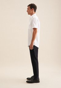 Witte korte mouwen shirt met twee borstzakken, gecombineerd met zwarte op maat gemaakte broek en zwarte schoenen, gezien vanaf de zijkant tegen een neutrale achtergrond.