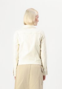 Veste en denim beige clair avec un design ajusté, col pointu et manches longues, présentant des détails de couture le long des coutures et un bas ourlé.