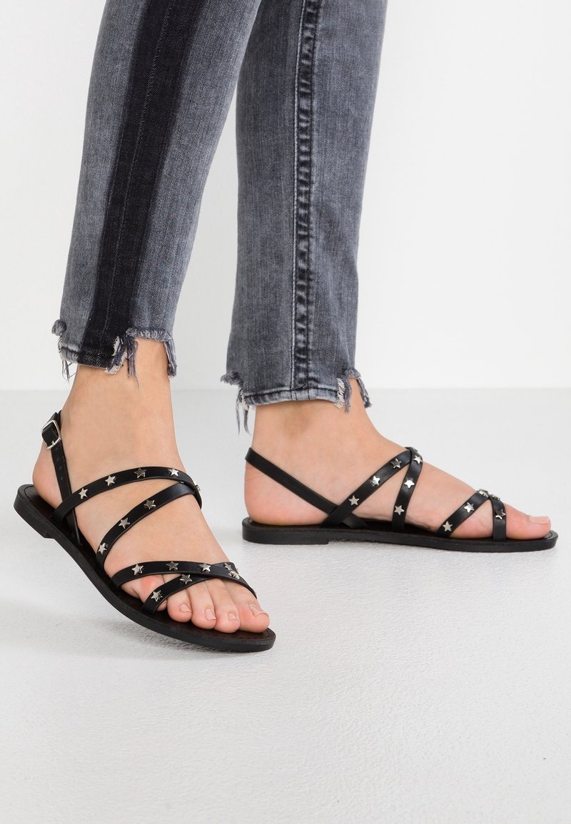 Pieds portant des sandales plates noires à brides avec des clous en forme d'étoile argentés, associées à un jean skinny gris à ourlet effiloché sur fond uni.