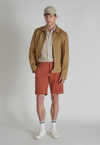Giacca leggera color tan, camicia beige, pantaloni corti color ruggine, sneakers bianche con calzini a strisce bianche e nere, cappello beige; outfit estivo casual.
