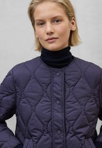 Quilted mørkelilla jakke med rund udskæring, to forlommer og knaplukning. Båret over en marineblå turtleneck sweater.