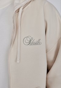 Beige Reißverschluss-Hoodie aus glattem Stoff, mit Kordelzugkapuze und gesticktem "Sitsilk"-Logo auf der Vorderseite.