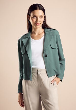Giacca teal con design colletto, un singolo bottone nero e due tasche frontali, abbinata a un top bianco e pantaloni beige.