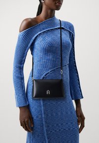 Donna che indossa un vestito aderente blu in maglia a coste con dettagli a spalle scoperte e una borsa a tracolla nera con chiusura in argento.