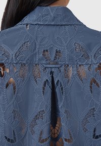 Blusa di pizzo blu con intricati ricami floreali, dettagli a taglio e colletto piegato. Etichetta metallica con marchio vicino al collo.
