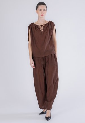 Joven mujer de pie, vistiendo una blusa marrón sin mangas de corte holgado con cuello con lazo y pantalones anchos a juego, zapatos negros puntiagudos.