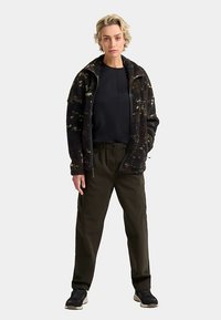 Black, textured fleece jacket with a high collar, zip closure, and golden speckled pattern; paired with olive green cargo pants and dark shoes.